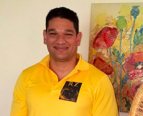 Elmer from Cuba is standing in front of a painting of flowers is smiling and wearing a yellow shirt.