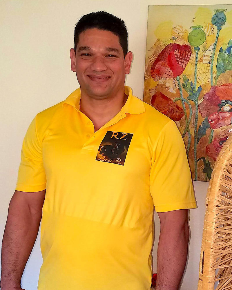 Elmer from Cuba is standing in front of a painting of flowers is smiling and wearing a yellow shirt.