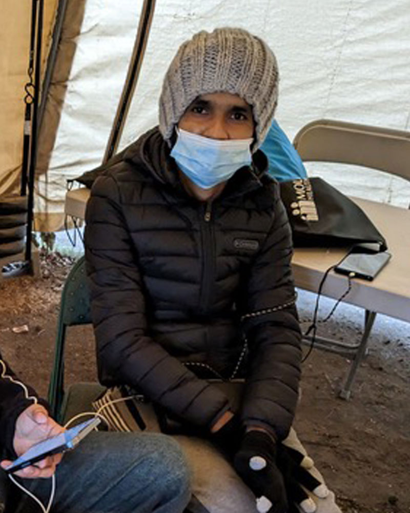Rama from Nepal is in a beanie and heavy jacket is sitting in an aid tent with a surgical mask on his face.