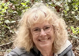 Aviva Furman, AIDNW welcome center volunteer - white, middle aged woman with blond curly hair and glasses. She is smiling widely.